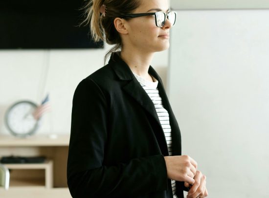 A confident woman wearing a black blazer and glasses in a modern office space.